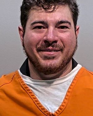 A man with short hair and a beard is smiling while wearing an orange prison jumpsuit.