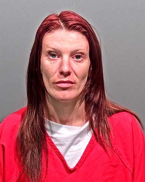 A woman with long red hair wearing a red jumpsuit is facing the camera in a mugshot setting.